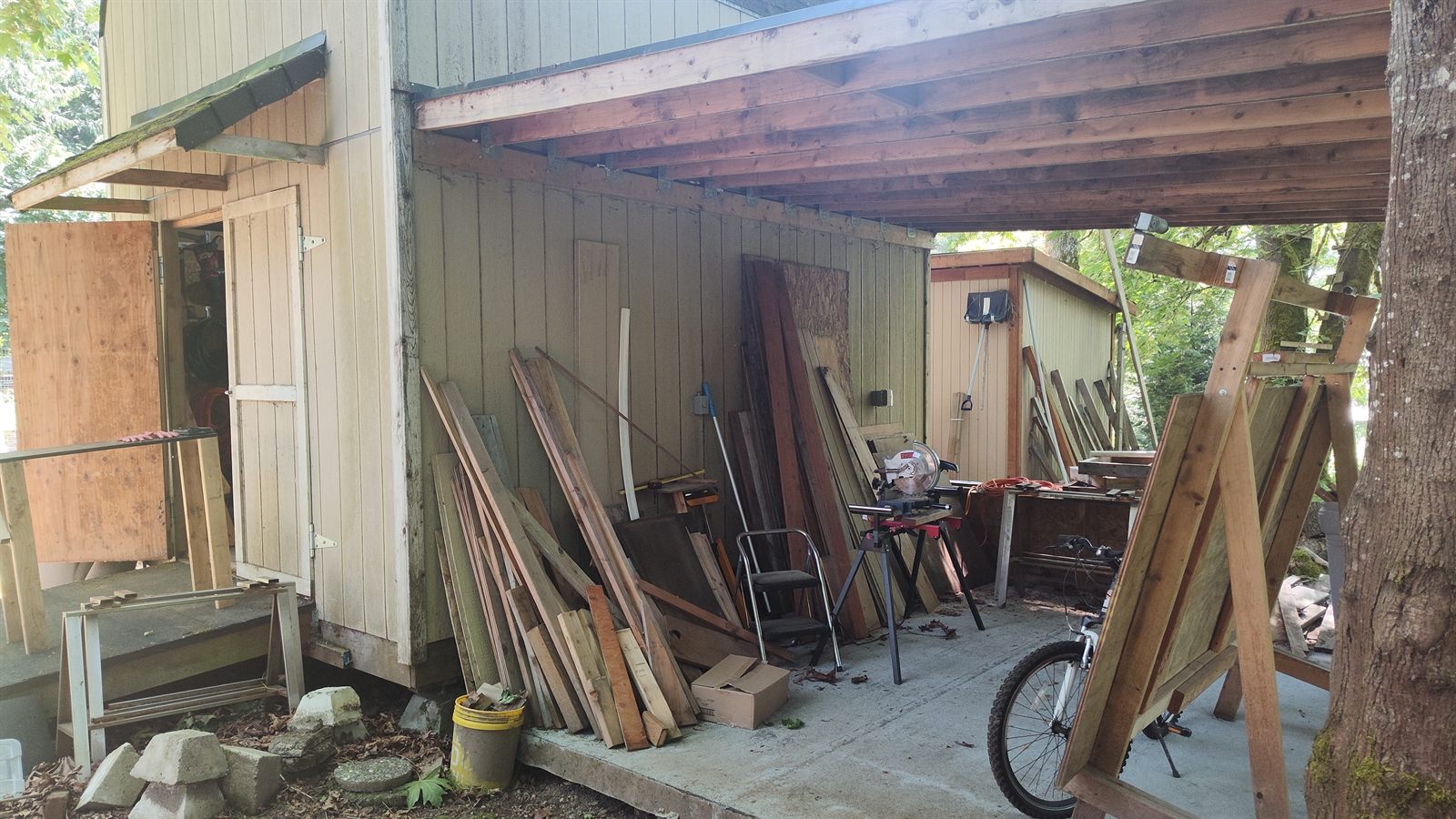 An outdoor covered work area with stored lumber leaning against the shop wall, a miter saw on a stand, and sawhorses